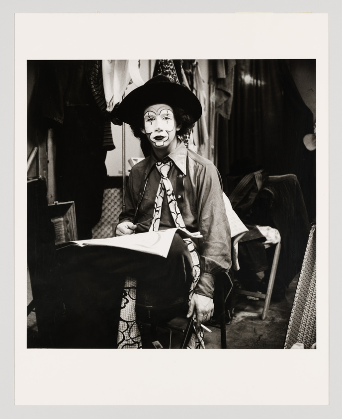 A black and white photograph of a person dressed as a clown, sitting and looking directly at the camera. The clown has a painted white face with exaggerated features, wearing a wide-brimmed hat, a patterned necktie, and a jacket. They are holding a piece of paper, possibly reading or drawing, in a backstage area with clothes hanging in the background.