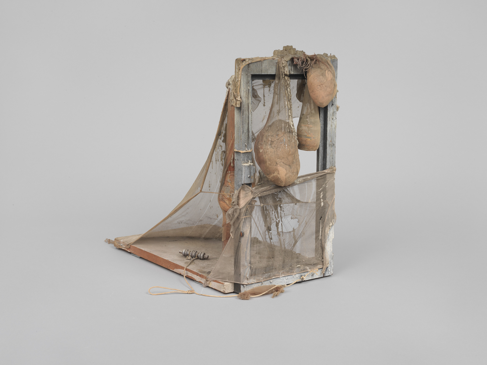 A worn and aged handmade model of a guillotine constructed from wood, fabric, and string, with two potato-like objects suspended where the blade would fall. The model is set against a plain, light-colored background.