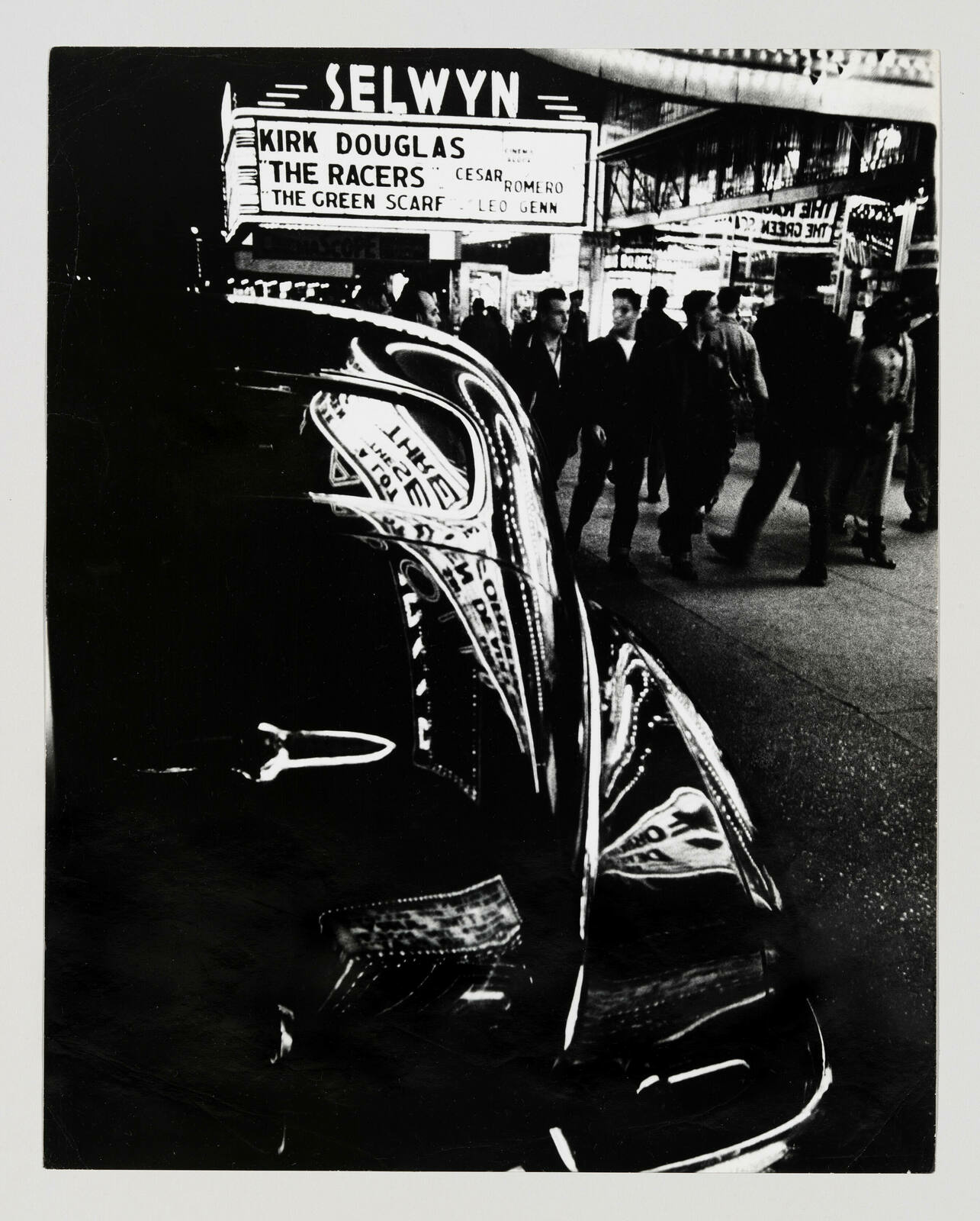 A black and white photograph capturing a busy city street scene with pedestrians walking past a movie theater marquee that reads "SELWYN" and advertises Kirk Douglas in "The Racers" and "The Green Scarf" with Cesar Romero and Leo Genn. The image is distorted and reflected on the shiny surface of a car, creating a surreal, fragmented effect.