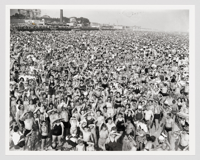 A densely packed crowd of beachgoers in vintage swimwear, with an amusement park visible in the background.