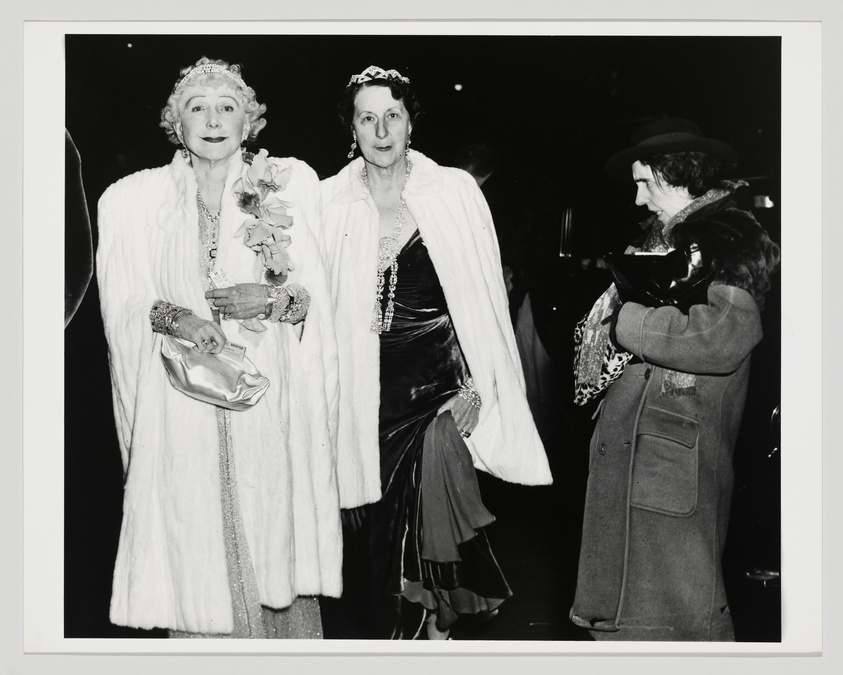 Two elegantly dressed women in fur coats and tiaras at a social event, with a third woman in a coat looking on.