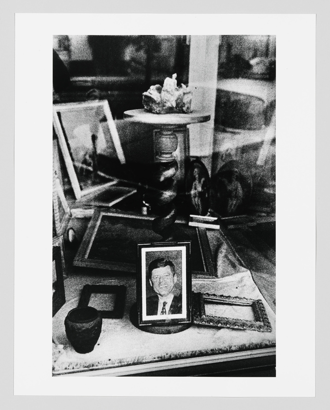 A black and white photo depicting a cluttered tabletop with various items including a framed portrait, an ashtray, a small urn, and several picture frames, some of which are empty. The scene has a reflective surface, suggesting it might be a glass table, and there's a blurred background that gives a sense of depth.