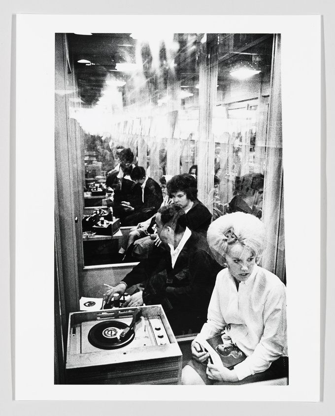 A black and white photograph capturing a moment inside a busy record listening booth. Several individuals are seen with headphones, engrossed in music. In the foreground, a woman with a beehive hairstyle sits next to a turntable, holding a record sleeve, while another person is changing a record. The reflection on the glass adds a layered, ghostly effect to the scene.