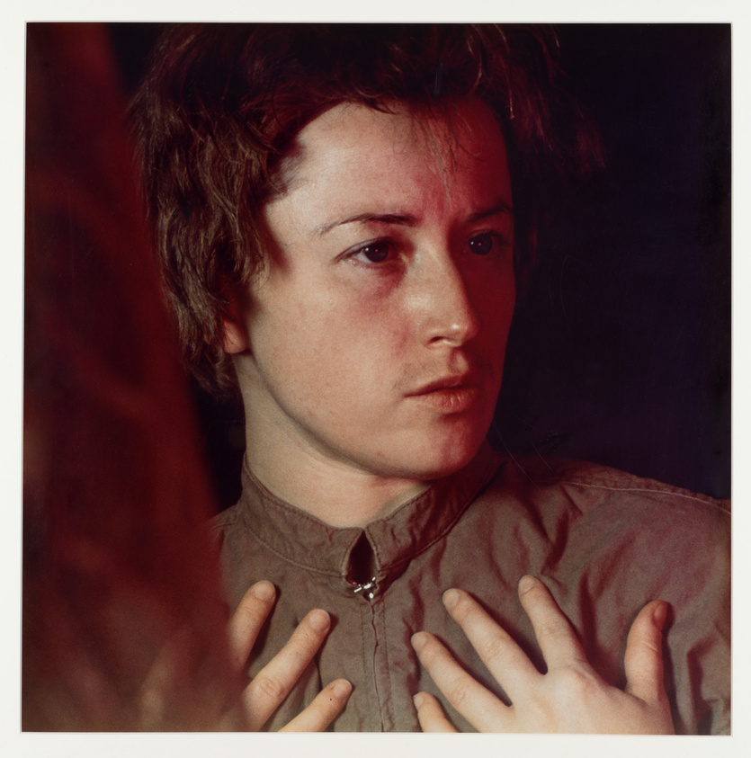 A contemplative individual with a neutral expression, hands gently placed on chest, against a dark background.