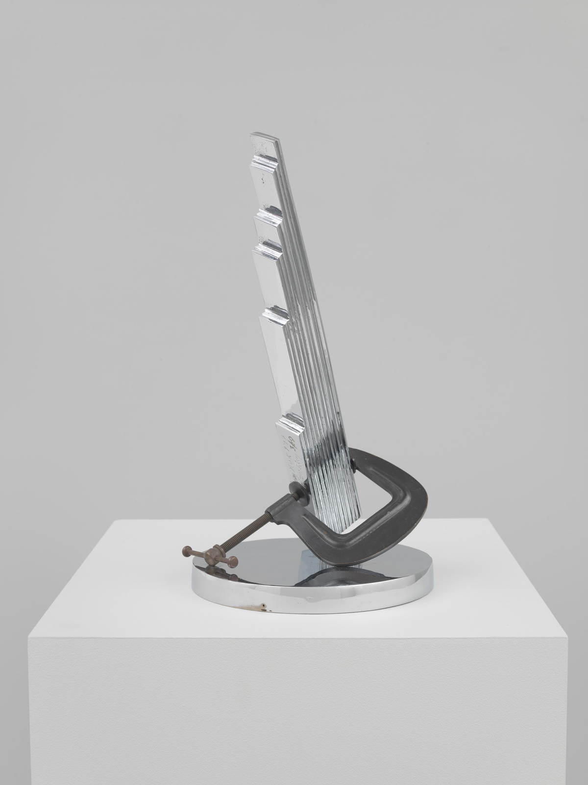 A metal sculpture clamped to a circular base, displayed on a white pedestal.