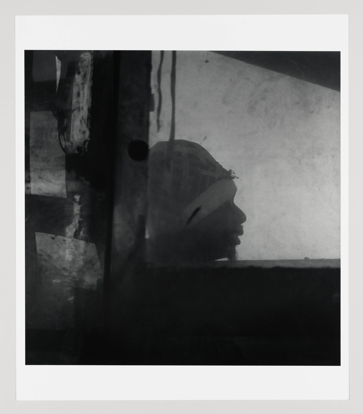 A monochromatic photograph capturing the silhouette of a person's profile through a hazy and textured windowpane, framed by the dark outlines of the window's structure.