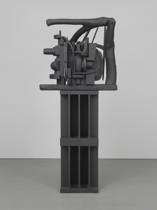 Dark gray abstract sculpture with mechanical elements on a tall pedestal against a plain background.