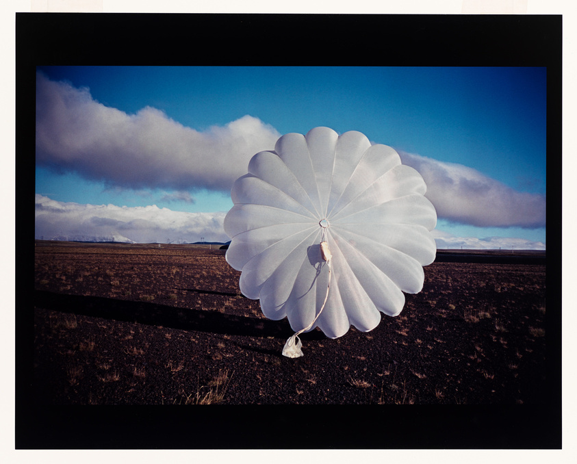 A large white circular parachute on a barren field with a blue sky and clouds in the background.