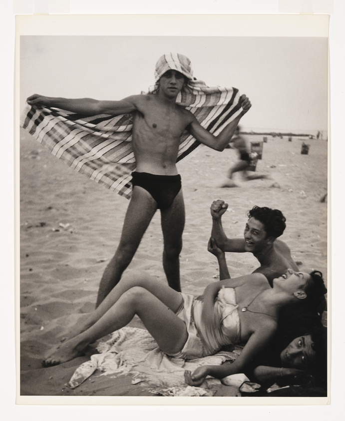 A black and white photograph capturing a candid moment on a beach. A shirtless man stands in the foreground, holding a striped towel draped over his head and outstretched arms, creating a makeshift cape. He wears dark swim trunks and a playful, determined expression. In the background, two women are lying on the sand, one propped up on her elbow looking up at the man with a smile, while the other lies on her back, seemingly laughing. The beach is populated with other beachgoers in the distance, and the sky appears overcast.