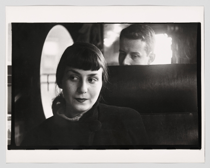 A black and white photograph capturing a woman in the foreground with a contemplative expression, looking slightly to the side. A man is partially visible in the background, seated behind her, facing the opposite direction. The setting appears to be a public space, possibly a train or a bus, with an arched window providing some light in the background. The image has a candid, vintage feel.