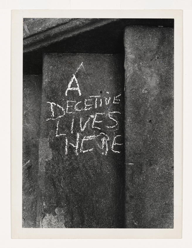 Black and white photo of a message "A DECEITVE LIVES HERE" scrawled on a wall.