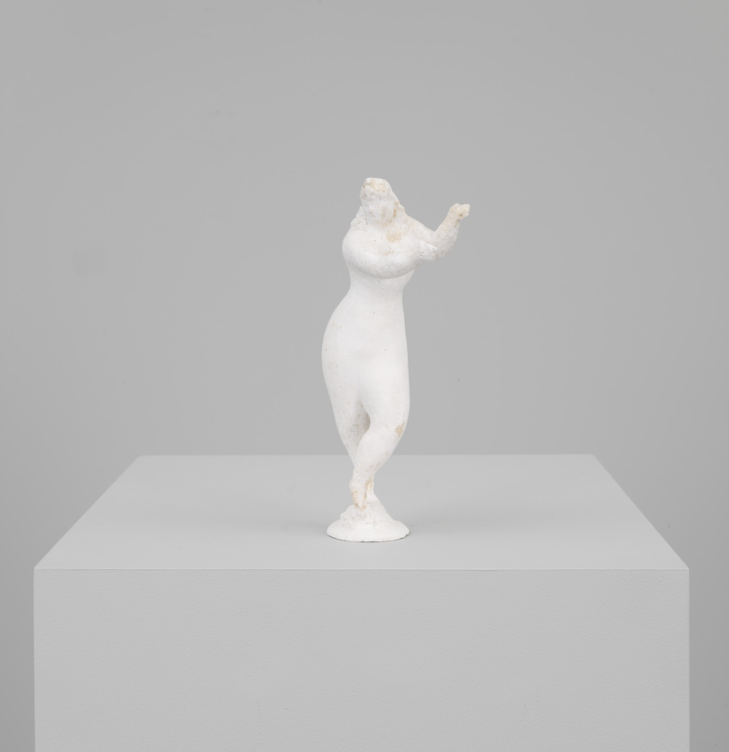 A white sculpture of a female figure with missing arms and head posed in a dancing position, displayed on a gray pedestal against a neutral background.