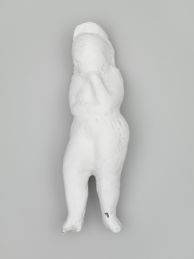 A white plaster sculpture of a human figure with no arms and indistinct facial features is mounted on a grey wall. The figure is in a standing position with its head tilted upwards and hands placed on the back of its head.