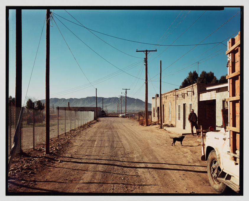 A dusty road stretches through a rural landscape with telephone poles lining one side. A dog walks across the road, and a person stands by a building to the right, next to a parked white pickup truck. In the background, a chain-link fence runs parallel to the road, and mountains rise under a clear blue sky.