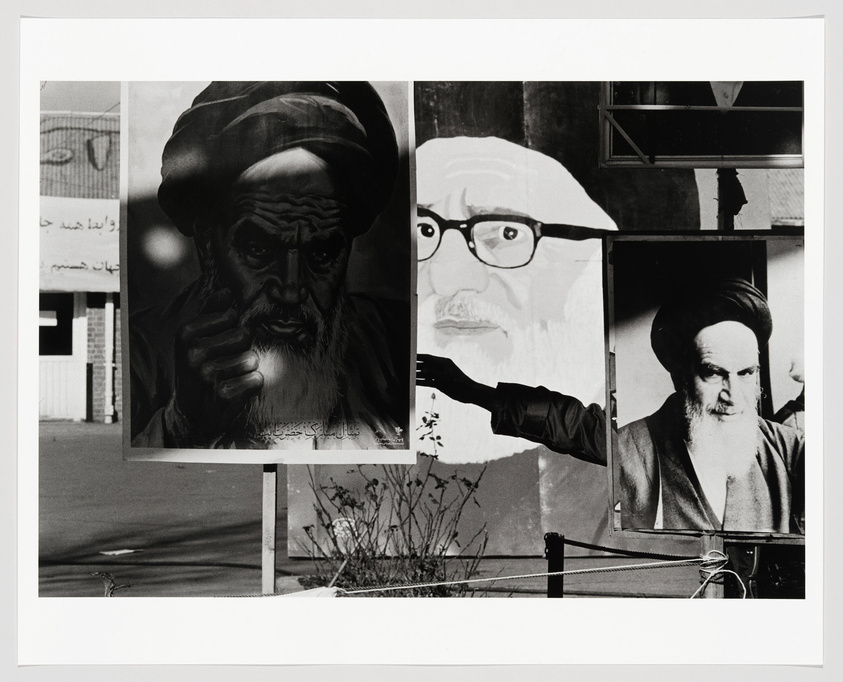 A black and white photograph showing three large posters of men with beards and traditional headwear, possibly religious or political figures, displayed outdoors. The left poster depicts a man pointing, the center poster features a man with glasses, and the right poster shows a man with a stern expression. Arabic script is visible on the building in the background.