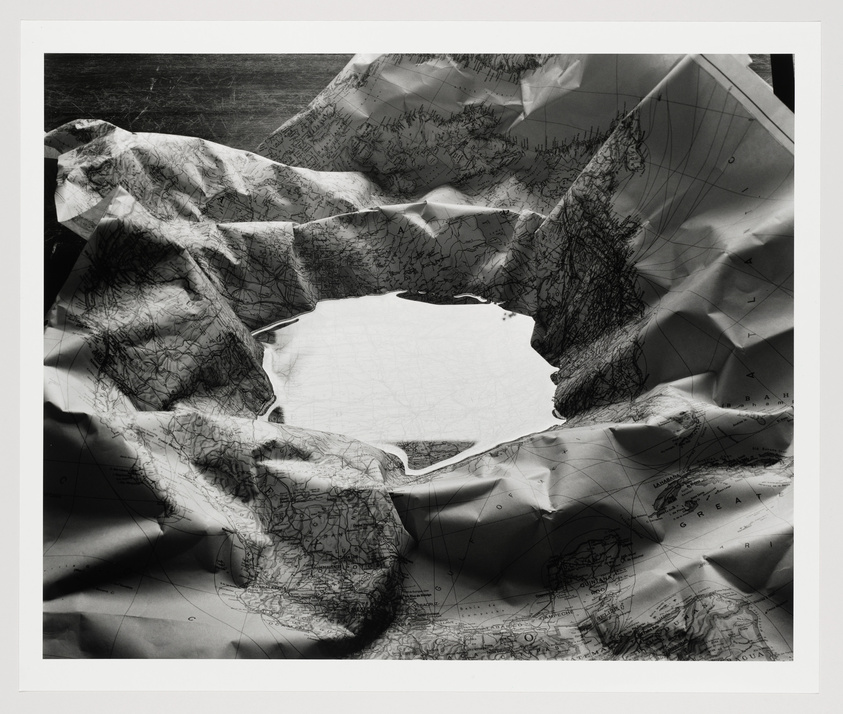 A black and white photograph of crumpled sheets of paper with map prints, featuring a central void where the paper has been torn away, revealing a blank space beneath. The image captures the intricate details of the map lines and the play of light and shadow across the textured surface.