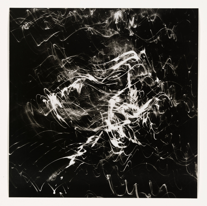 A dynamic black and white abstract photograph featuring a chaotic pattern of white light streaks against a dark background, resembling a long exposure of moving lights.