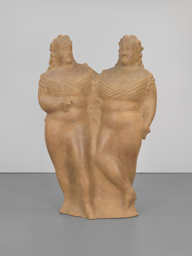 A sculpture of a conjoined twin figure with two heads and a single body, featuring intricate patterns across the chest, displayed in a gallery with a white wall and grey floor.