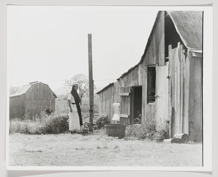 A black and white photograph depicting a woman standing outside a rustic wooden barn with another building visible in the background. The woman is dressed in a long dress with a light-colored apron and her hair is pulled back. In the foreground, there's tall grass and a metal tub, with various items leaning against the barn's exterior.