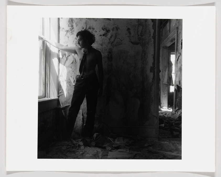 A black and white photograph capturing a person standing in a dilapidated room with peeling walls, reaching out towards a window with tattered curtains. The room is filled with debris and the light from the window casts a dramatic contrast of shadows and highlights across the scene.