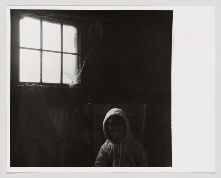A black and white photograph depicting a young child sitting in a dimly lit room, wearing a hooded sweatshirt. The child is looking downward with a contemplative or somber expression. Light streams in through a window with a visible cobweb, casting a bright rectangle on the opposite wall and partially illuminating the child's face. The room appears aged, with peeling wallpaper and a draped cloth or garment hanging to the left. The image conveys a sense of stillness and introspection.