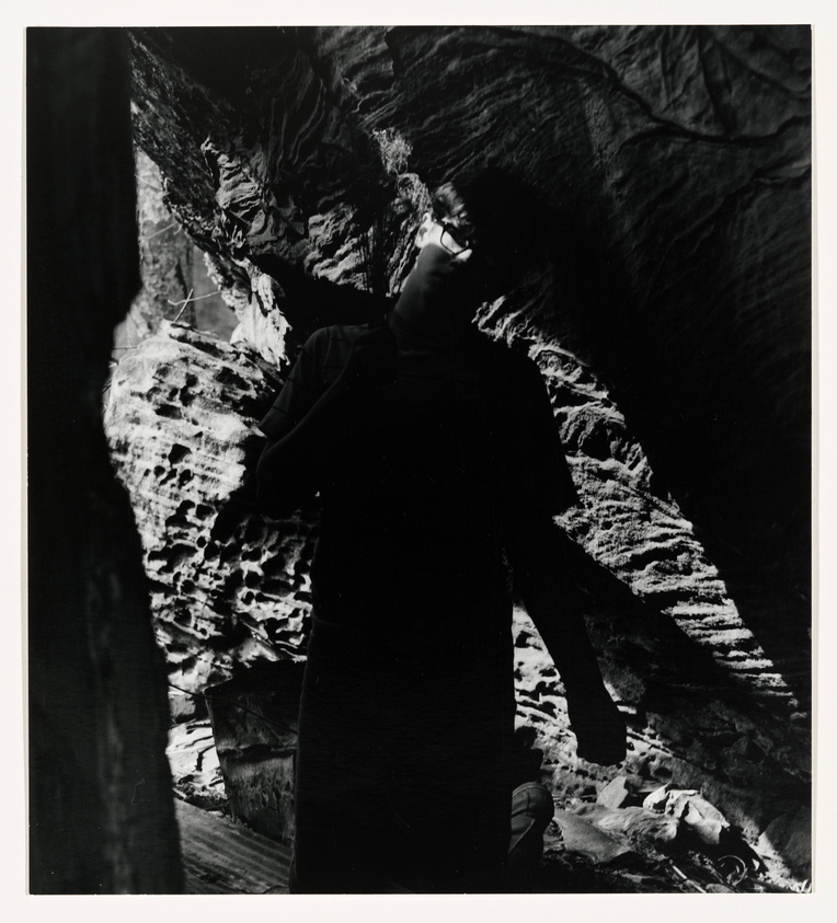 A black and white photograph capturing a person in a dark, textured cave or rocky area. The individual is dressed in dark clothing and appears to be looking upwards towards a source of light that illuminates their face and the surrounding rock formations. The contrast between light and shadow creates a dramatic and moody atmosphere.