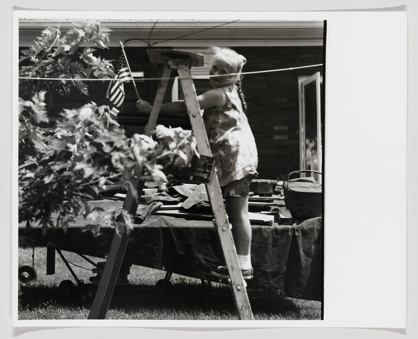 A black and white photograph capturing a moment in a residential backyard where a child is climbing a ladder. The child is wearing a dress and has their back to the camera. In the background, an American flag is visible in a window, and various items such as a wheelbarrow and a bucket are scattered around the yard, suggesting outdoor activities or chores. The image has a vintage feel, with its monochrome tones and the composition giving a glimpse into a casual, domestic scene.