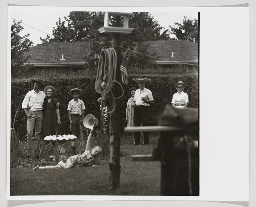 A black and white photograph depicting a group of people standing in a garden. On the left, a man and a woman wearing hats stand with two boys, one also in a hat, and all are looking towards the camera. On the right, two women and a man stand with their hands clasped in front, also facing the camera. In the foreground, a maypole with ribbons and a doll lying on the ground can be seen, suggesting a festive or celebratory event. The image has a vintage feel, with the clothing styles and garden setting evoking a mid-20th-century ambiance.