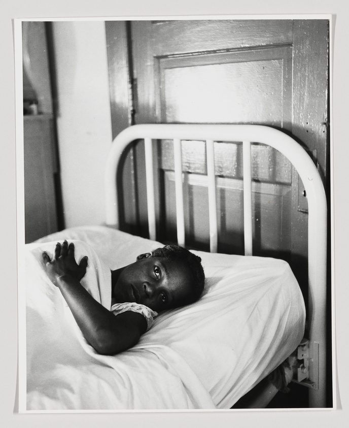 A black and white photograph of a young child lying in a hospital bed, looking directly at the camera with a poignant expression. The bed has a white metal frame and is positioned against a wall with a wooden door. The child is partially covered by a white sheet, and the room appears to be vintage or from an earlier era.