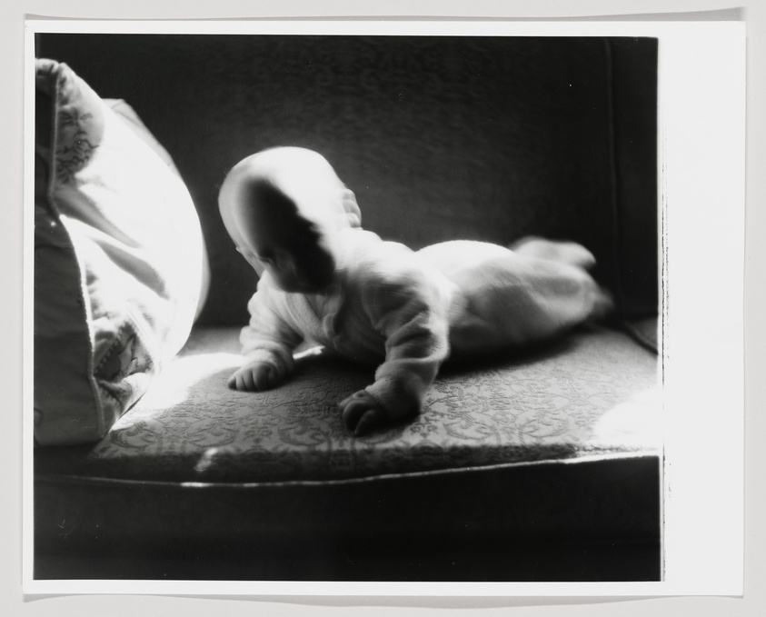 A black and white photograph capturing a baby lying on its stomach on a patterned surface, with a strong light source coming from the left, creating a high-contrast image with distinct shadows.