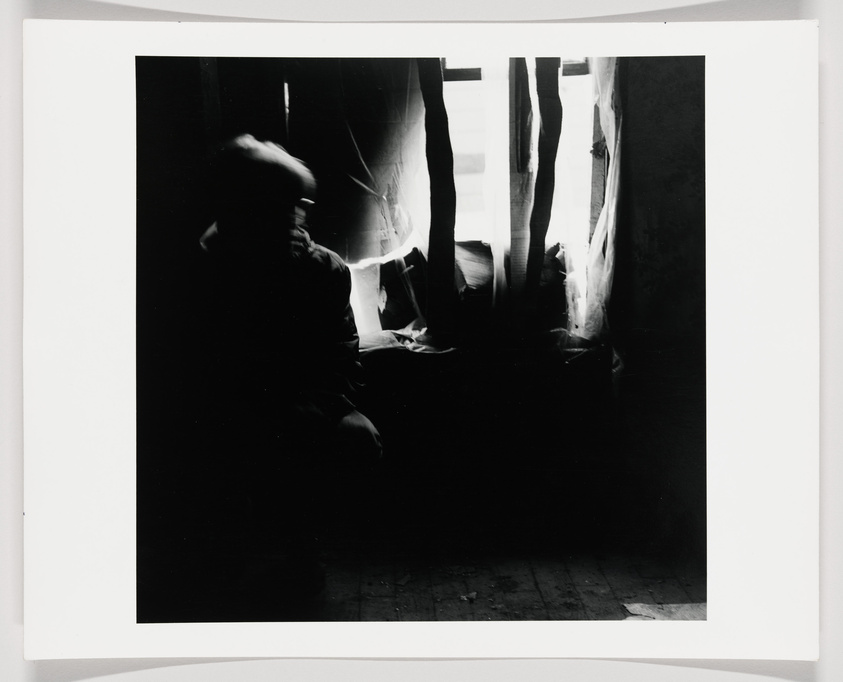 A black and white photograph featuring a blurred silhouette of a person sitting to the left with light streaming through a window with tattered curtains on the right, creating a stark contrast between light and shadow. The image has a vintage, eerie atmosphere and is bordered by a white frame.