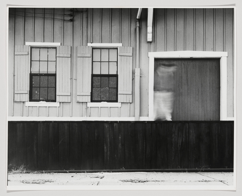 Black and white photograph of a wooden building with two windows with shutters on the left and a blurred curtain behind a glassless window frame on the right, above a dark wooden fence.