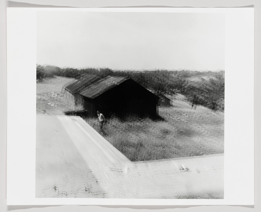 A black and white photograph depicting a rural scene with a weathered wooden barn on the right and a person walking along a road to the left of the barn. The landscape includes open fields and sparse trees in the background under a light sky.