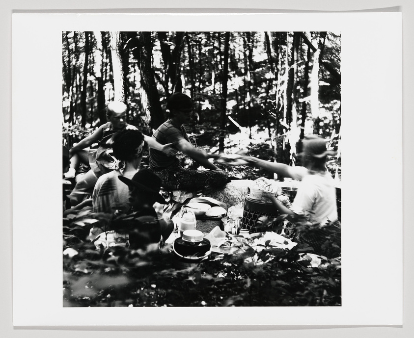 A black and white photograph of a group of people enjoying a picnic in a forested area, with dappled sunlight filtering through the trees. Some individuals are seated on the ground while others are reaching out across a picnic blanket laid out with various dishes and utensils. The focus is soft, adding a nostalgic or timeless quality to the scene.