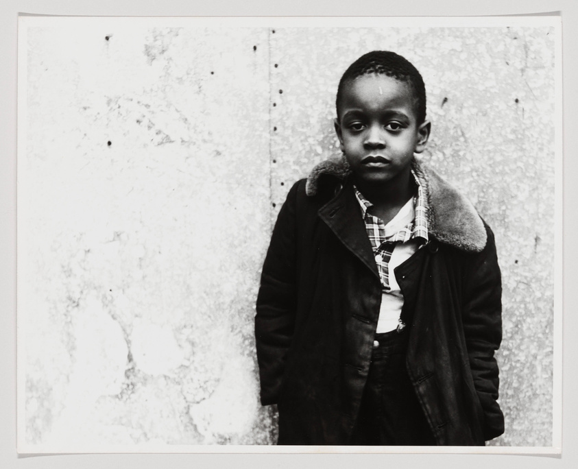 A black and white photograph of a young boy standing in front of a textured wall, wearing a large coat with a fur-lined collar over a plaid shirt. The boy has a solemn expression on his face and is looking directly at the camera.