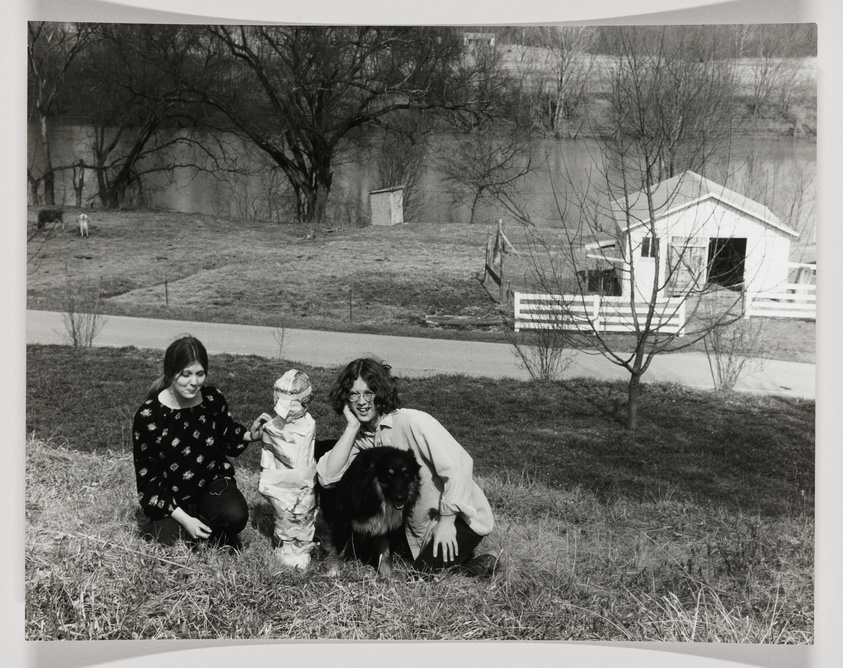 Two women and a child are crouching on a grassy hillside with a black dog, smiling towards the camera. In the background, there's a white shed with a fence around it, a leafless tree, and a river with trees on the far bank. The setting appears to be in a rural area.