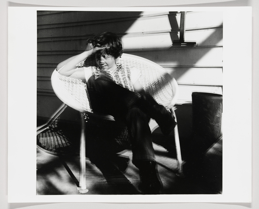 A black and white photograph of a person sitting relaxed in a white, round wicker chair on a sunny porch, shielding their eyes from the sun with one hand. Shadows from the chair's pattern are cast on the deck, and a dark container is visible in the corner.