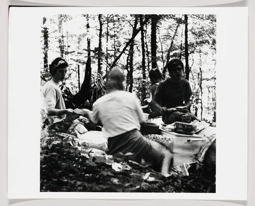 A black and white photo of a group of people enjoying a picnic in a forested area, with trees in the background and picnic items spread out on a cloth.