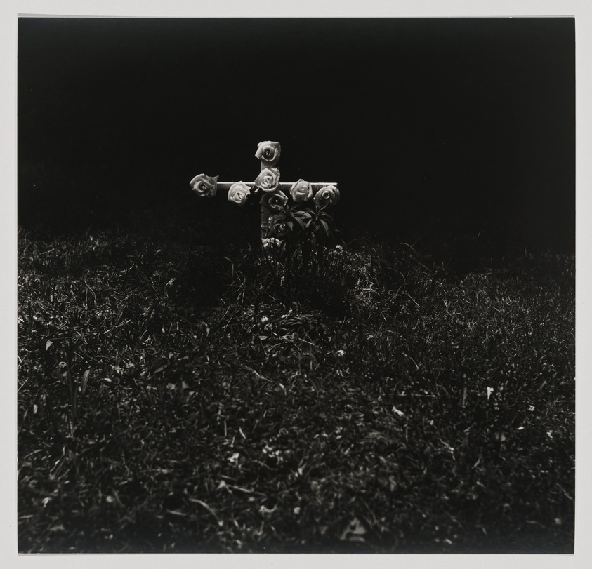 A black and white photograph depicting a small cross adorned with roses, standing in a grassy area with a dark, featureless background.