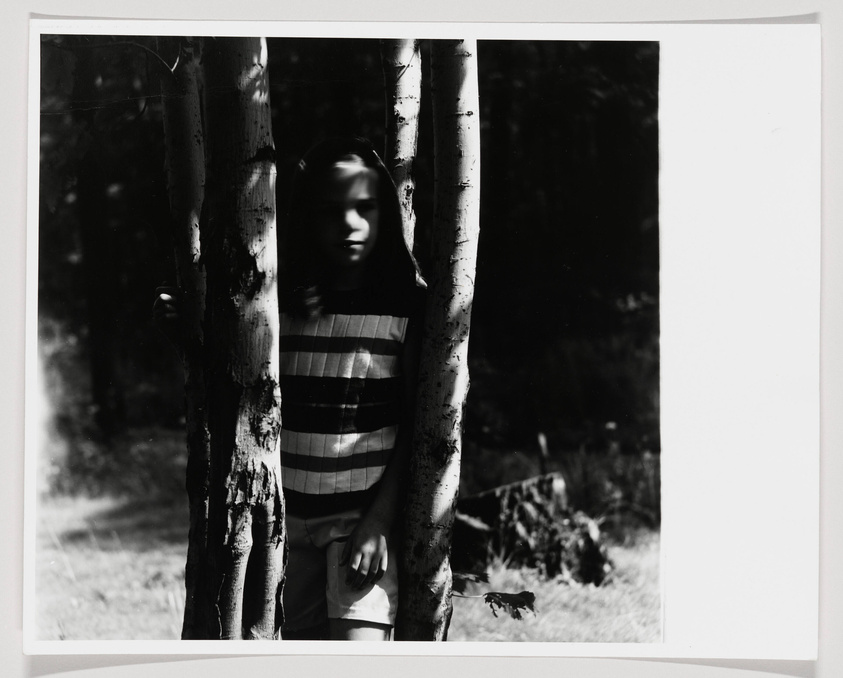 A black and white photograph showing a person partially obscured by tree trunks, wearing a striped shirt and standing in a wooded area. The image has a high contrast with some areas overexposed, creating a ghostly effect on the person's face.