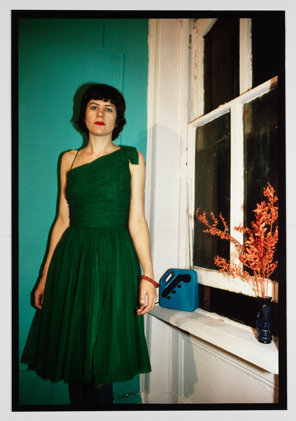 A woman in a one-shoulder green dress stands next to a window with peeling white paint. The wall behind her is painted in a contrasting turquoise color. To her right, there is a small blue radio on the floor and a vase with red foliage on the windowsill. The image has a candid, unposed quality.