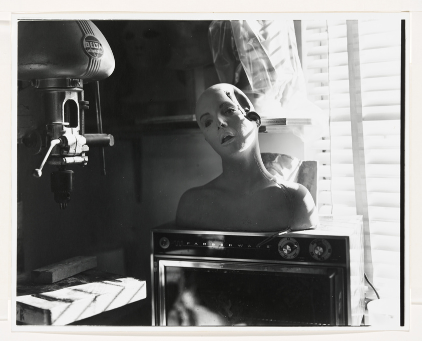 A black and white photograph featuring a bald mannequin head and torso placed on a microwave oven, with a drill press visible on the left. The scene is lit with high contrast, casting sharp shadows on the wall and the microwave. The background includes partially closed blinds and a draped cloth, contributing to a surreal and somewhat eerie atmosphere.