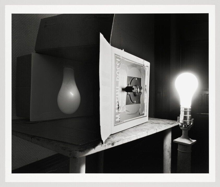 A black and white photograph showing a creative setup with a light bulb theme. On the left, a large white light bulb is suspended inside a dark box, creating a silhouette effect. In the center, a smaller light bulb is mounted on a stand inside a microwave oven with its door open, suggesting an experiment or artistic display. On the right, a glowing light bulb is screwed into a simple socket on a stand, illuminating the scene. The background is nondescript, focusing attention on the light bulbs and the microwave.