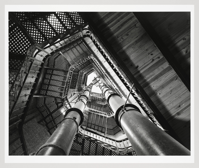 A black and white photograph looking up the interior of a spiral staircase with intricate metalwork and a wooden ceiling, framed by two large vertical pipes.