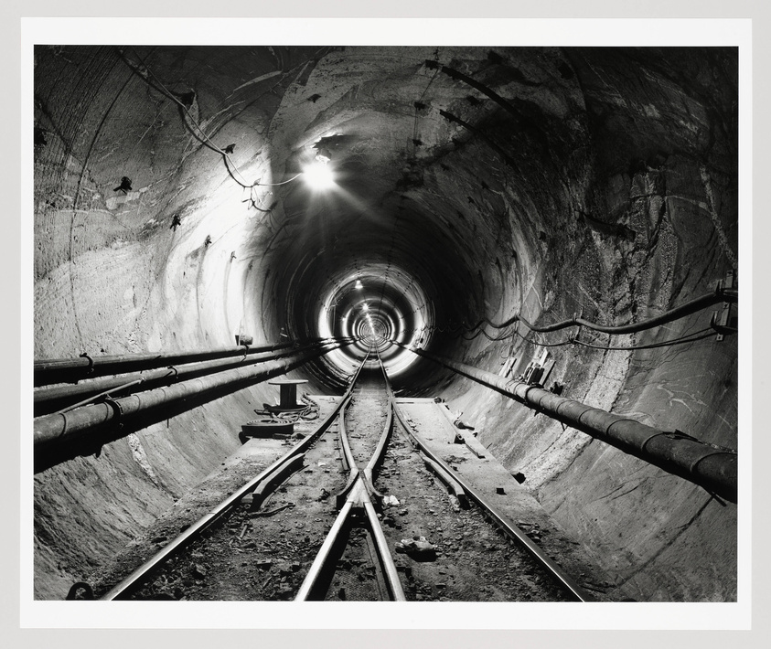A black and white photograph of a railway tunnel with tracks leading into the distance. The tunnel is illuminated by a bright light at the far end and various smaller lights along the curved walls. Pipes and cables run alongside the tracks, and the rough texture of the tunnel walls is visible.