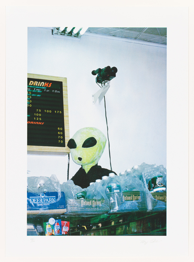 Person in alien mask behind a water bottle display with a drinks menu board in the background.