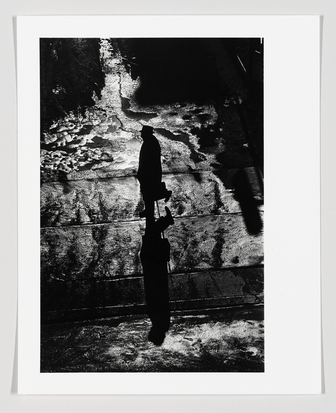A high-contrast black and white photograph depicting the silhouette of a person walking on a textured surface with the shadow of the figure prominently displayed. The light reflects off the ground, creating a pattern that contrasts with the dark figure and shadow.