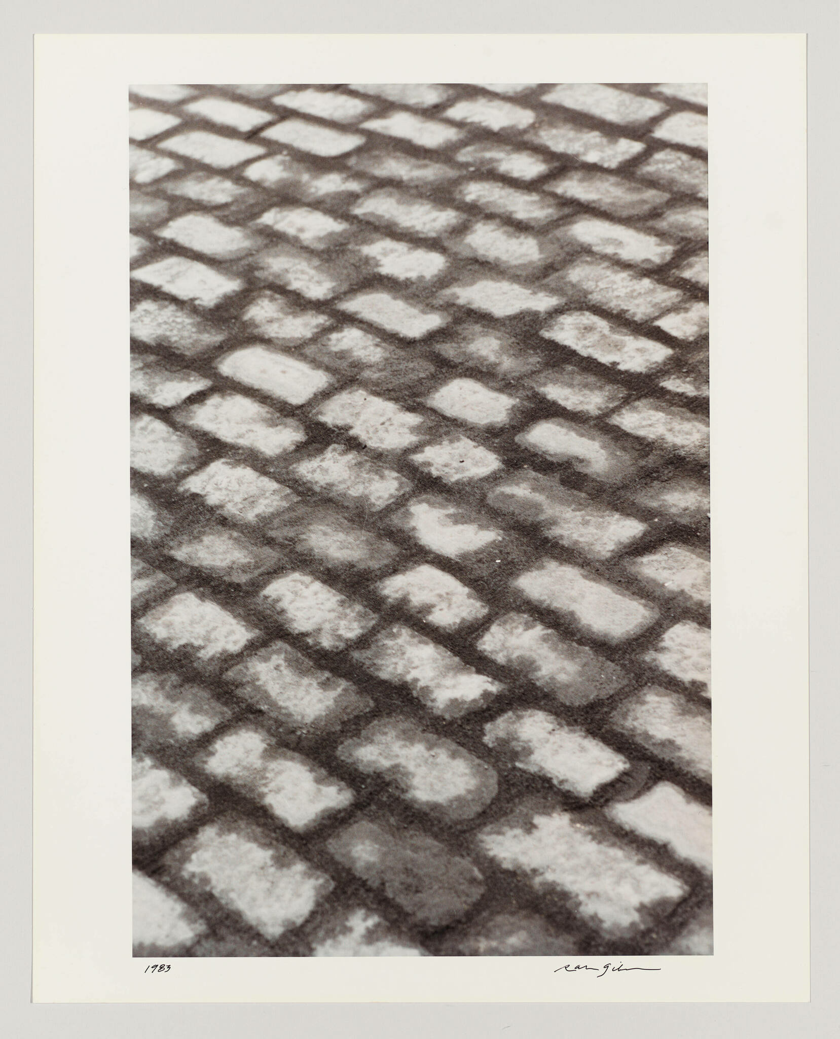 A monochrome artwork depicting a repetitive pattern of cobblestones with contrasting light and dark tones, suggesting the play of light and shadow. The image is framed with a white border, and there are handwritten signatures and numbers at the bottom, indicating it's a limited edition piece.