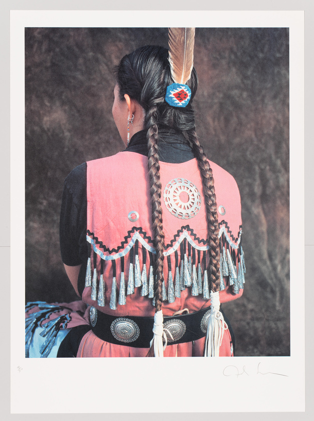 Woman in traditional Native American attire viewed from behind, showcasing intricate beadwork and a feathered hairpiece.