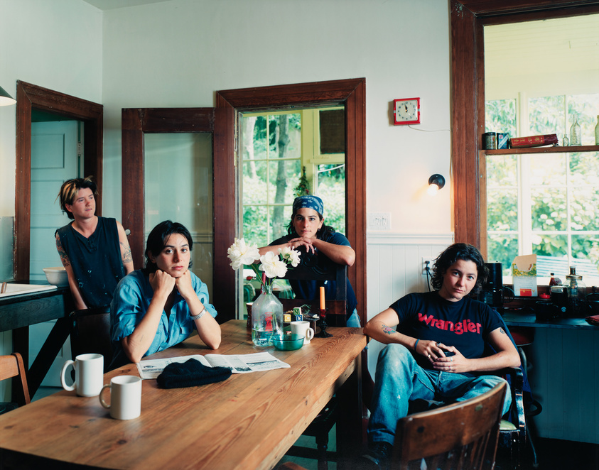 Four individuals are seated around a wooden dining table in a cozy kitchen with white walls and wooden trim. The person on the left is leaning against the wall, looking to the side, while the next person is seated at the table with their chin resting on their hands, gazing directly at the camera. The third individual, sitting across the table, is leaning forward with their elbows on the table and hands clasped, also looking at the camera. The fourth person is seated at the end of the table with a relaxed posture, legs crossed, and looking slightly away from the camera. The table has a few items on it, including a newspaper, a vase with white flowers, and two white mugs. The kitchen has a homey feel with visible shelves and a window that lets in natural light.